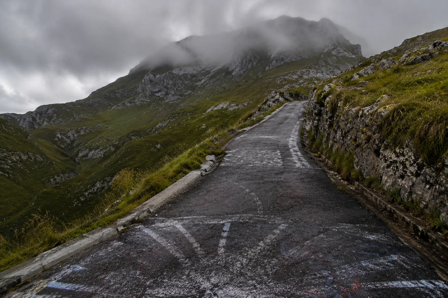 Steep mountain road with painted gradient markings climbing through mist on the Alto de l'Angliru in Asturias